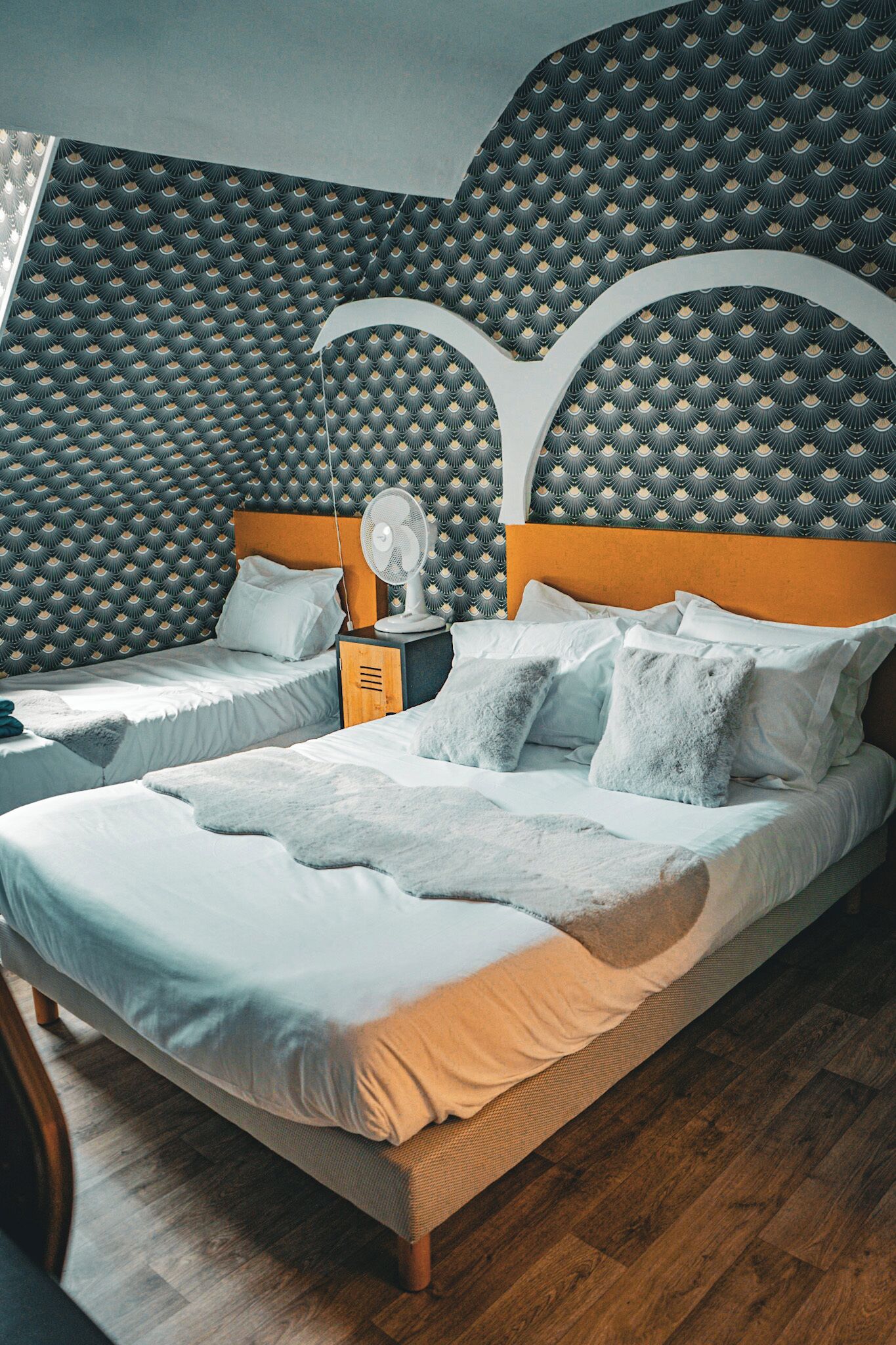 Ein Schlafzimmer mit zwei Betten, einem hölzernen Kopfende, einem Nachttisch mit Ventilator und gemustertem Wandpapier.