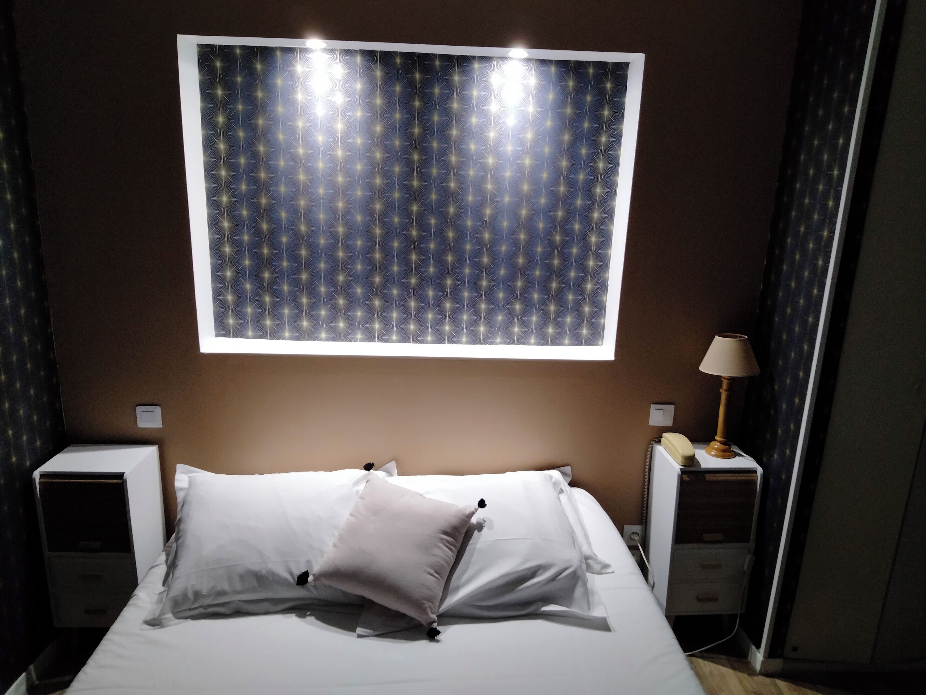 Ein Hotelzimmer mit einem Bett, zwei Nachttischen, einer Lampe und einem Fenster mit Sternenmuster.