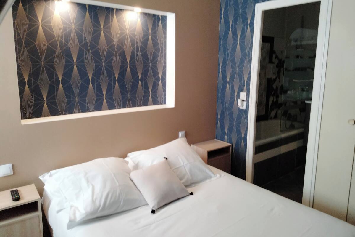 Una habitación de hotel con cama, almohadas blancas, una mesita de noche, control remoto y un espejo.