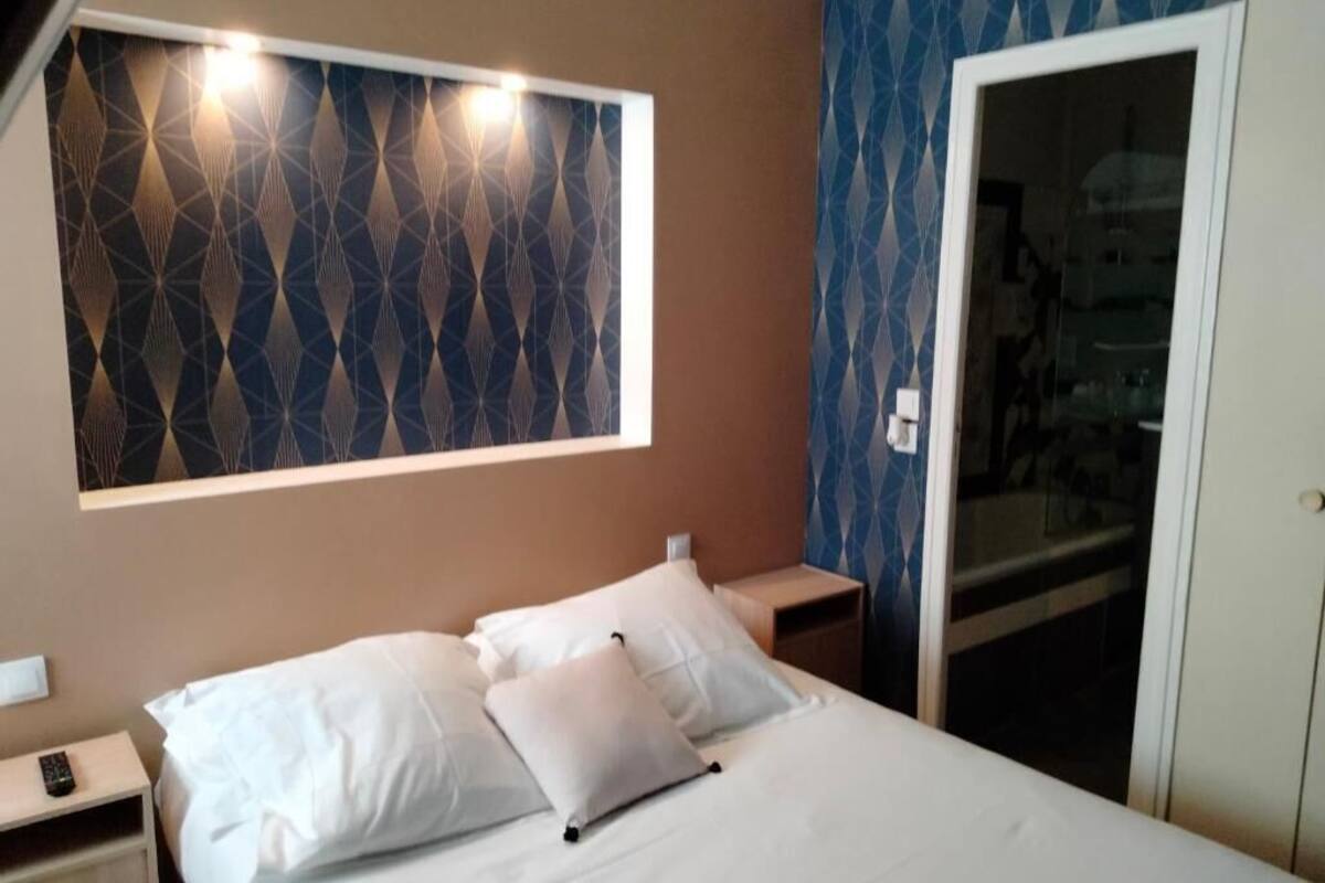 Habitación de hotel con cama, mesita de noche, lámpara de pared y puerta que da al baño.