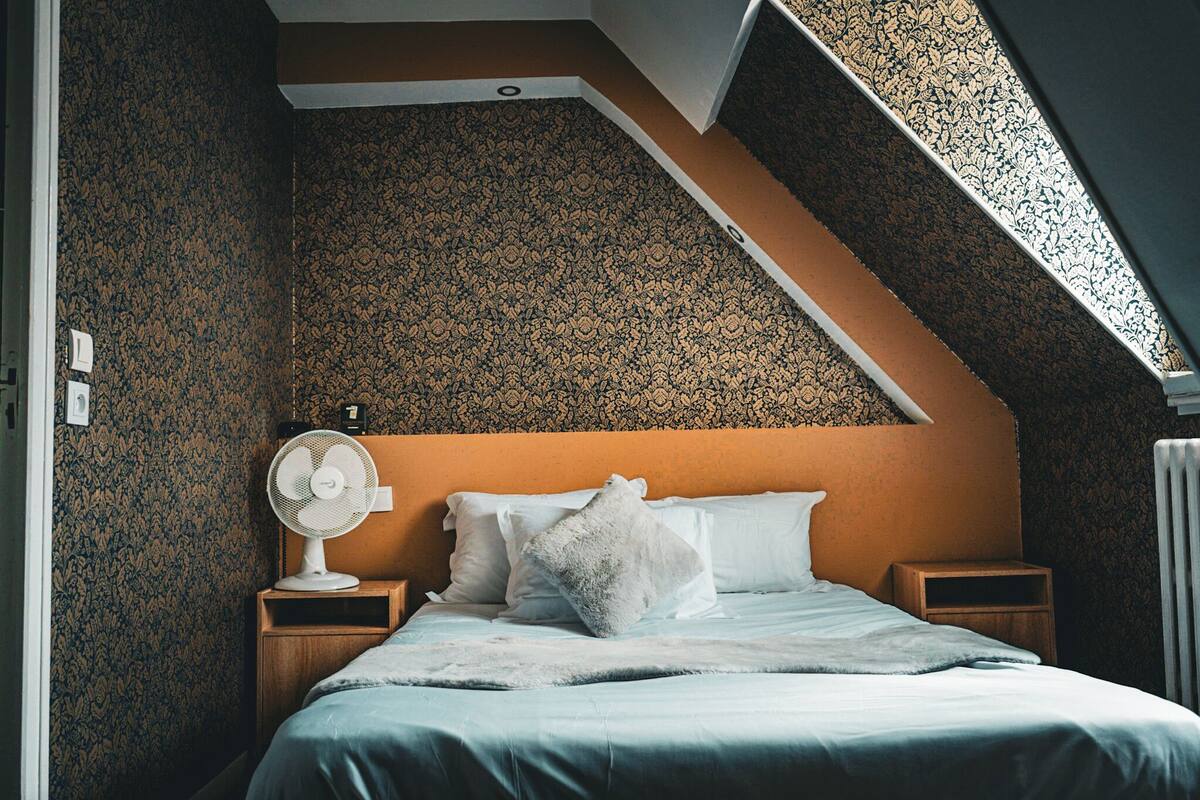 Un dormitorio con techo inclinado, papel tapiz estampado, cama con ropa de cama blanca, ventilador blanco y una mesita de noche de madera.