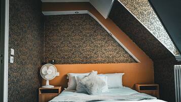 Ein Schlafzimmer mit schrägem Dach, Muster-Tapete, einem Bett mit weißer Bettwäsche, einem weißen Ventilator und einem kleinen Nachttisch aus Holz.