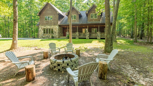 Un lodge con camino in pietra, poltrone Adirondack e un braciere in pietra in una zona boscosa.