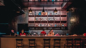 A bar area with multiple shelves of bottles, a counter with bar stools, and a person in a red shirt behind the bar. - Ban Ban Hotel (Phnom Penh)