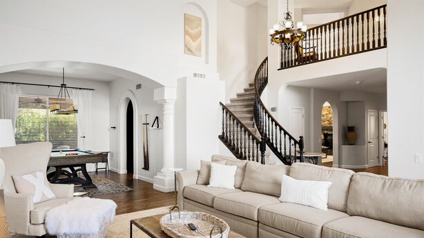En rummelig stue med en stor beige sofa, et sofabord og et spiseområde med et billardbord. En trappe med sort gelænder fører op til en etage.