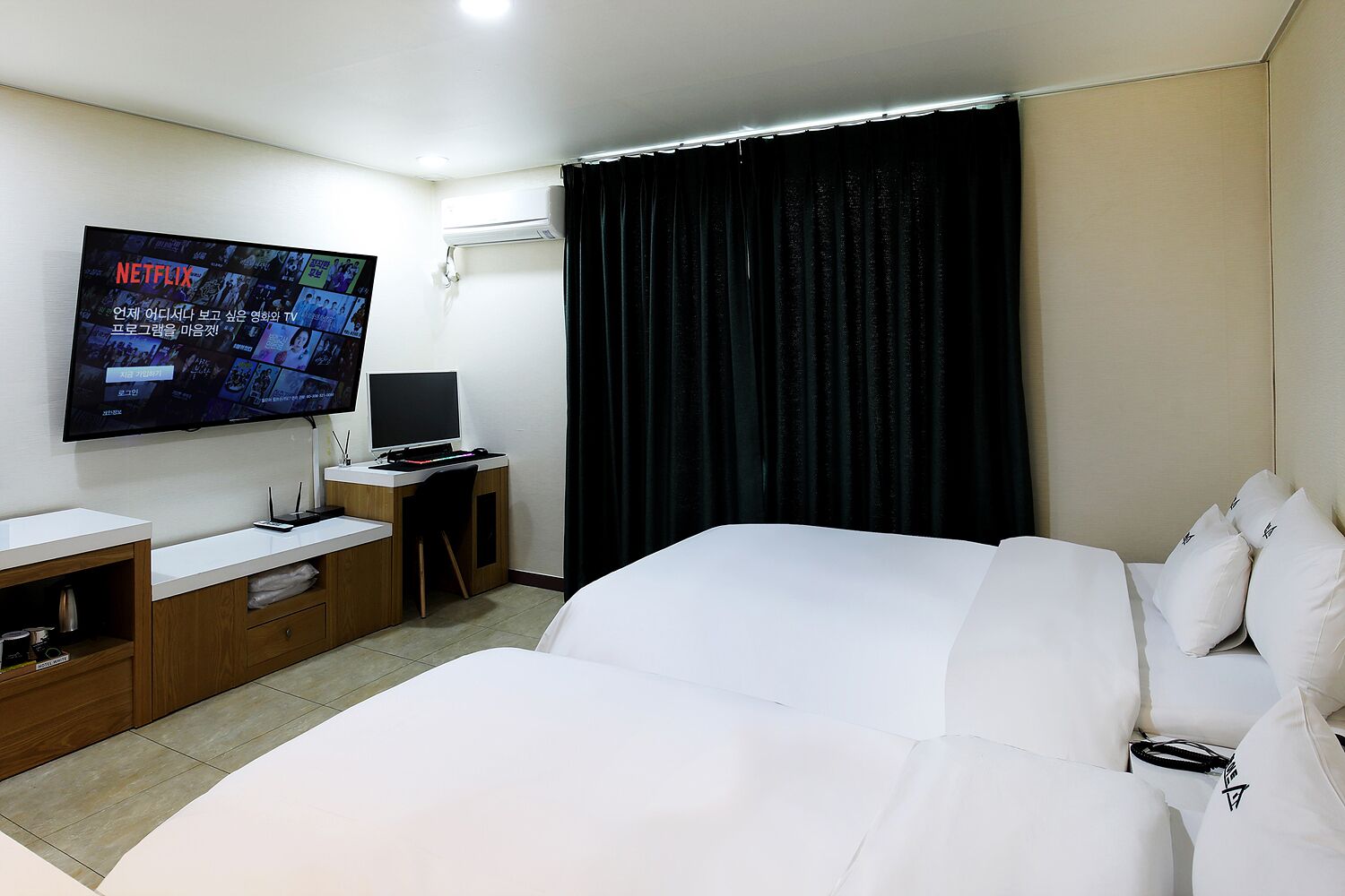 A hotel room with a large bed, a desk with a computer, a flat-screen TV mounted on the wall, and a window with dark curtains.