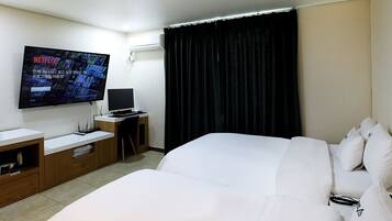 A hotel room with a large bed, a desk with a computer, a flat-screen TV mounted on the wall, and a window with dark curtains.