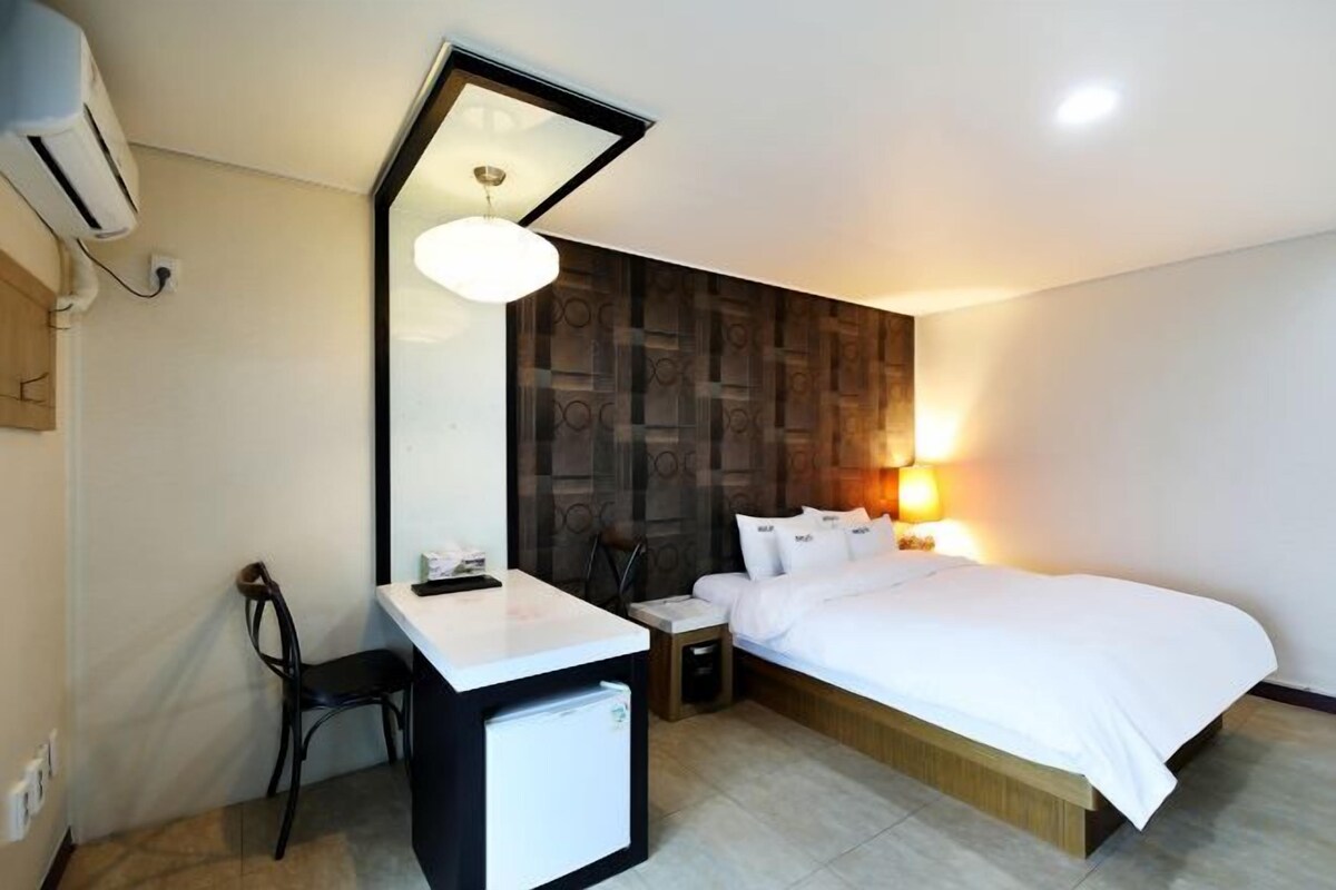 A hotel room with a bed, a desk, a chair, a bedside table, and a wall-mounted air conditioner.