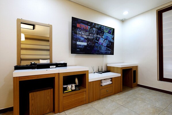 A modern bathroom with a wooden vanity, a large mirror, a flat-screen TV mounted on the wall, and a window with blinds.