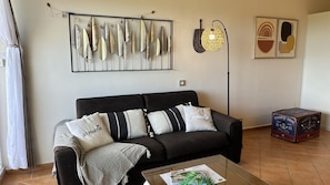 A living room with a black sofa, patterned cushions, a glass coffee table, a framed feather artwork, a geometric light fixture, and a vintage Pacific Soul box.