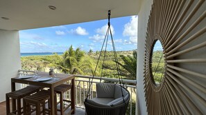 A balcony with a wooden table set for dining, a hanging chair, and a view of a tropical landscape with palm trees and a beach.