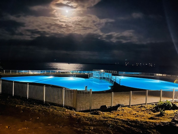 A large, illuminated swimming pool at night with a view of the ocean and a bridge in the background.