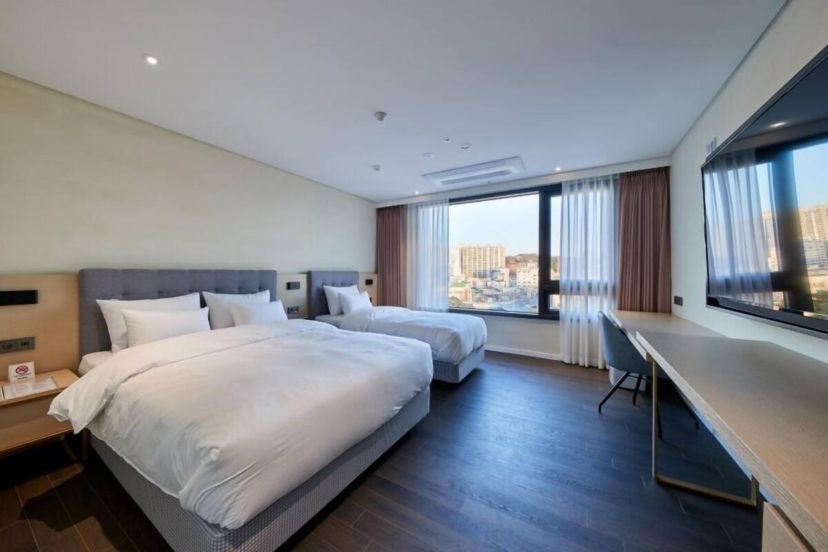 A hotel room with two beds, a desk, and a large window offering a city view.