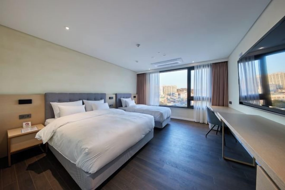 Habitación de hotel con dos camas, un escritorio y un ventanal grande con vistas a la ciudad.