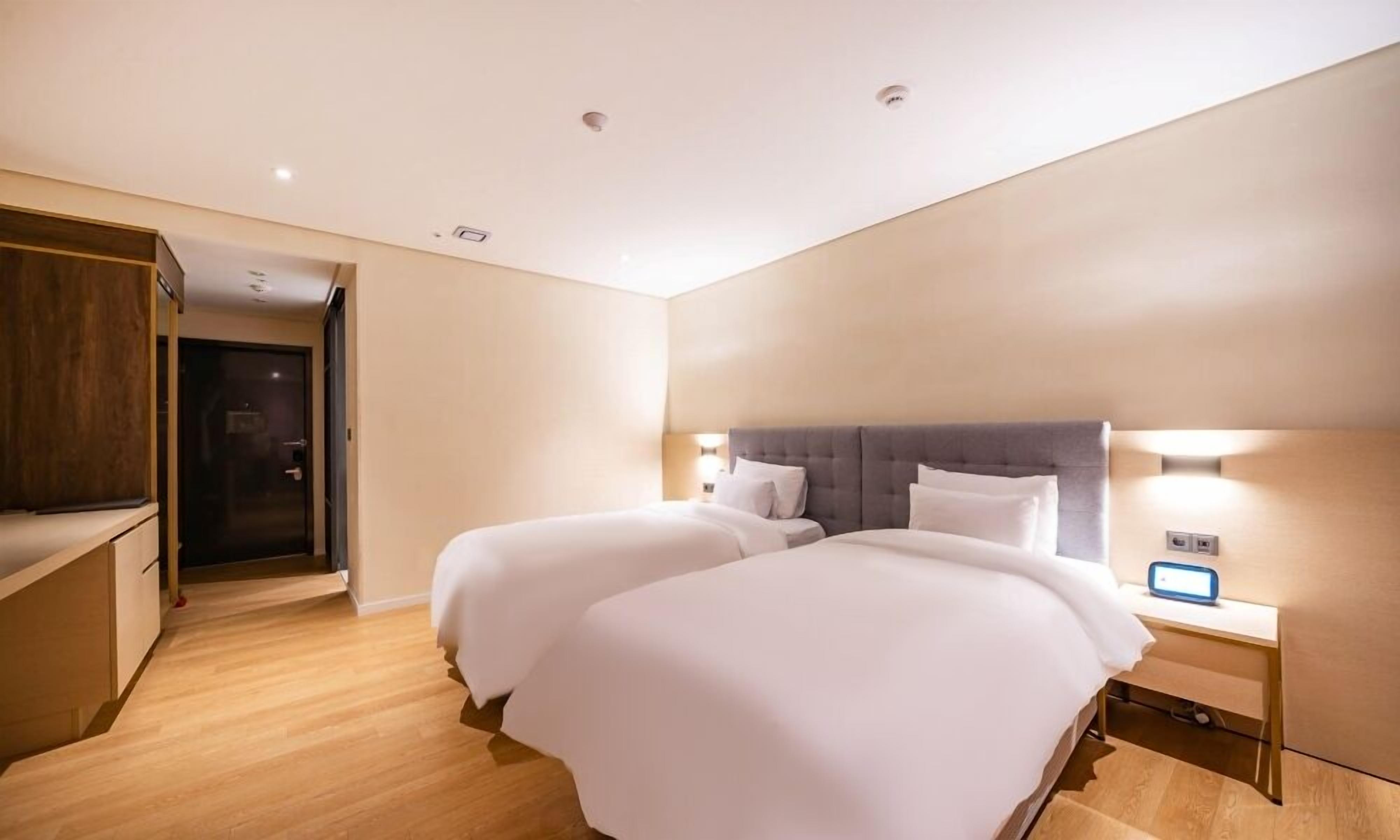 A hotel room with two beds, wooden flooring, a wardrobe, and a bedside table with a lamp.
