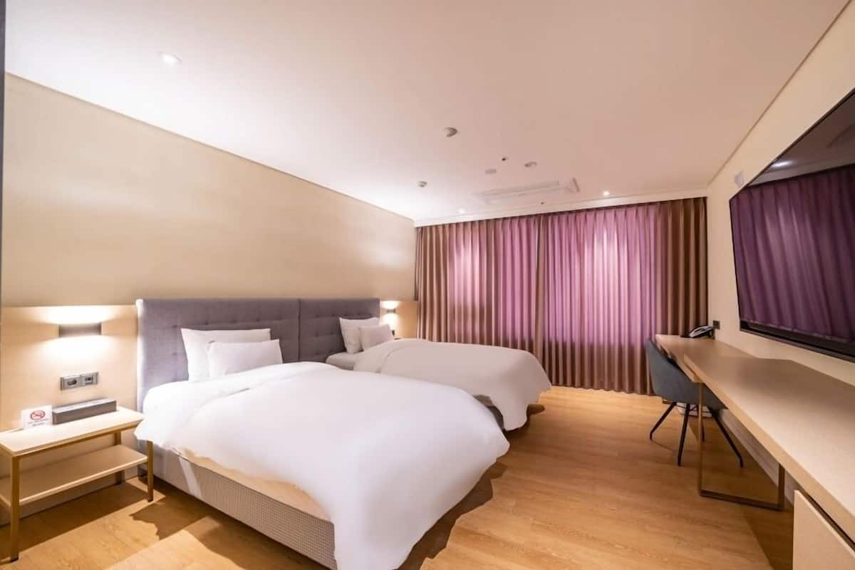 A hotel room with two beds, a desk, a flat-screen TV, and a large window with curtains.