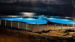 Una piscina grande e iluminada de noche, con vista al océano y un puente al fondo.