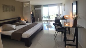 Habitación de hotel con cama, escritorio, silla, planta en maceta y balcón con vistas.
