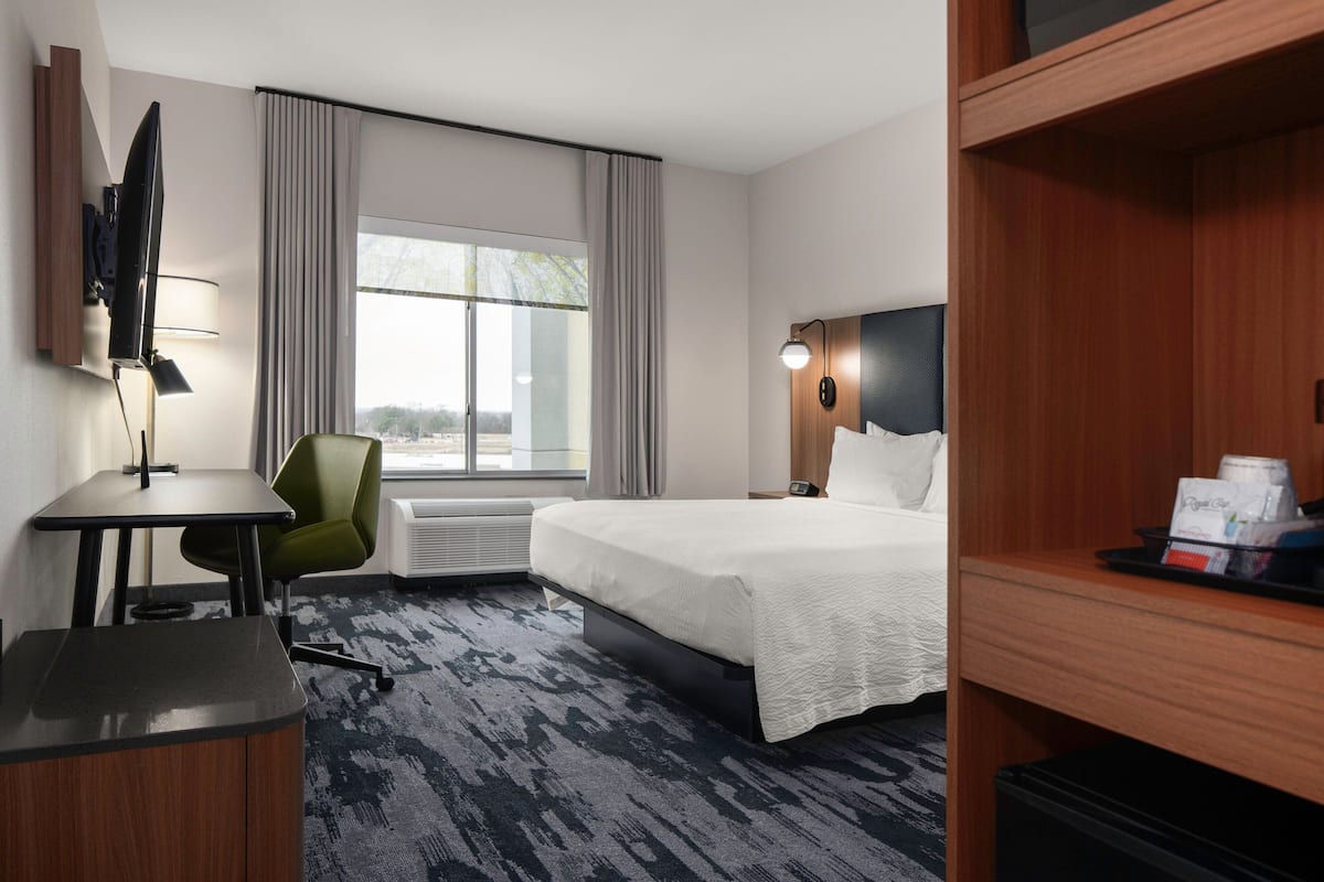 A hotel room with a bed, a desk with a chair, a TV mounted on the wall, and a window with curtains.