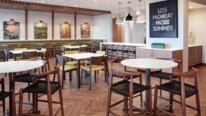 Interior moderno de restaurante con suelo de madera, mesas redondas y sillas. Se observan obras de arte en la pared y un letrero que dice "LESS MONDAY MORE SUMMER".