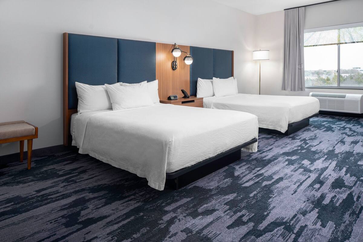 A hotel room with two beds, a bedside table, a lamp, and a large window.