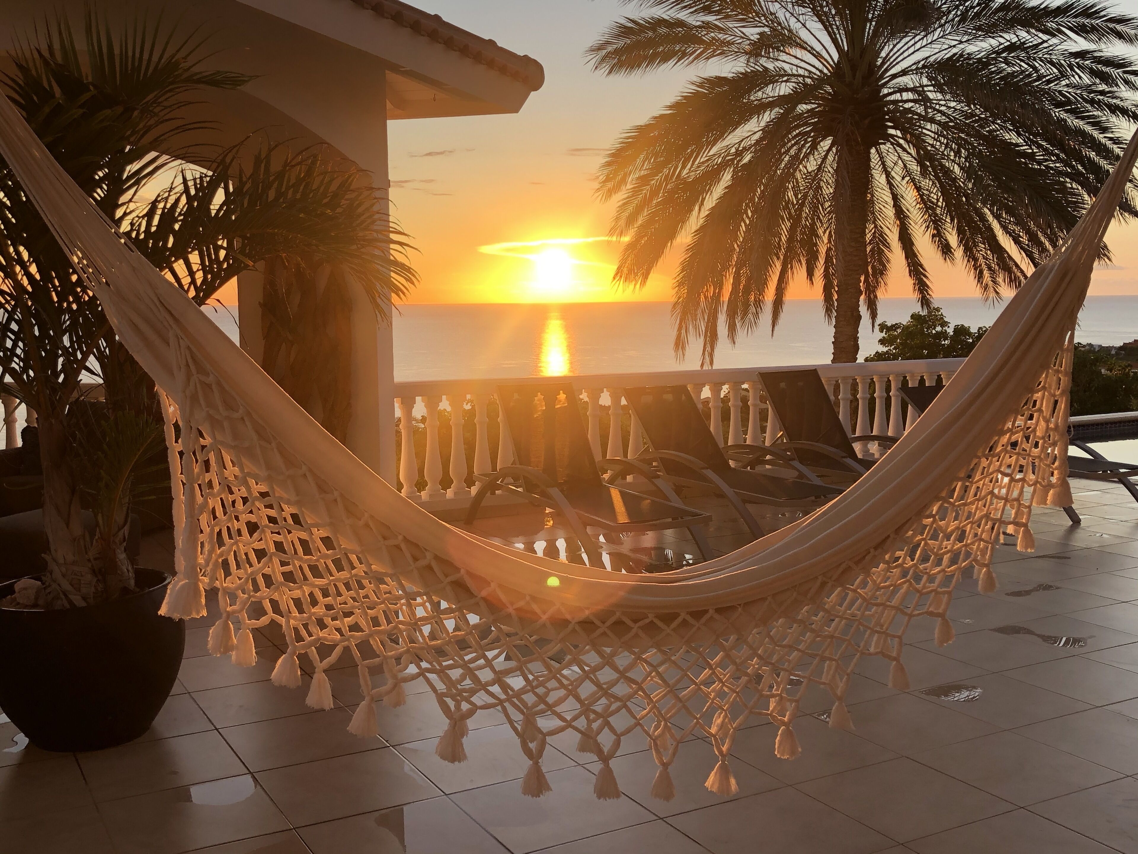 Un'amaca su un patio piastrellato con vista sul tramonto sull'oceano, palme e un parapetto.