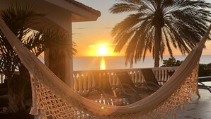 Un hamac sur une terrasse carrelée, d’où l’on peut admirer le coucher de soleil sur l’océan, les palmiers et une balustrade.