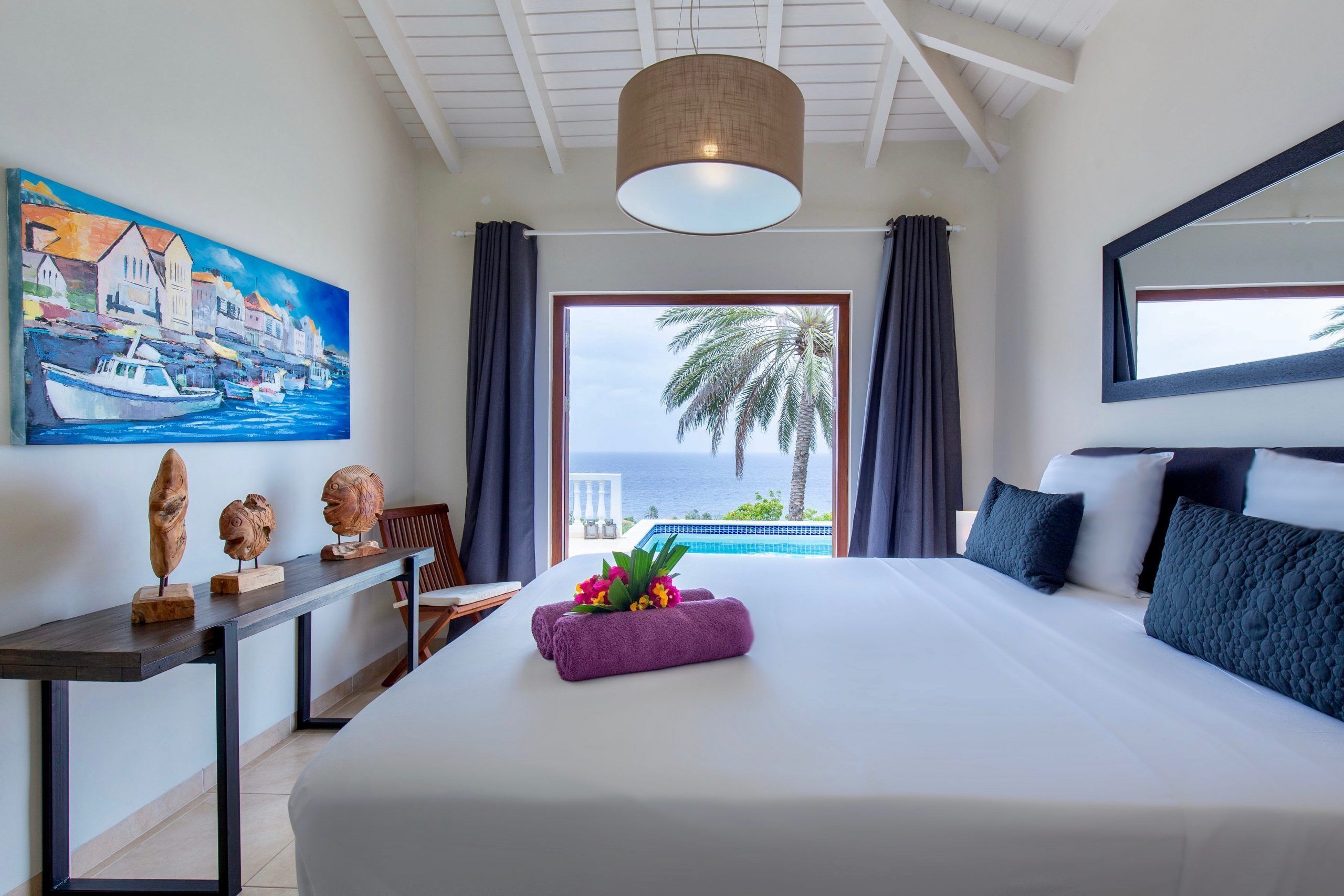 Camera d'albergo con un letto grande, un quadro raffigurante una scena costiera, vista sul mare e una palma all'esterno.