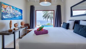Une chambre d’hôtel avec un grand lit, un tableau représentant un paysage côtier, une vue sur la mer et un palmier à l’extérieur.