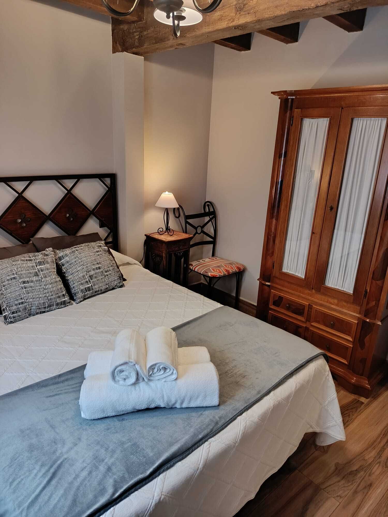 Una cama bien hecha con una colcha gris y toallas enrolladas, un armario de madera con cortinas, una mesita con una lámpara y una silla con un cojín.