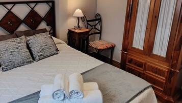 Una cama bien hecha con una colcha gris y toallas enrolladas, un armario de madera con cortinas, una mesita con una lámpara y una silla con un cojín.