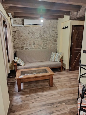 A room with a stone wall, wooden floor, a sofa with patterned cushions, a wooden coffee table, and a door.