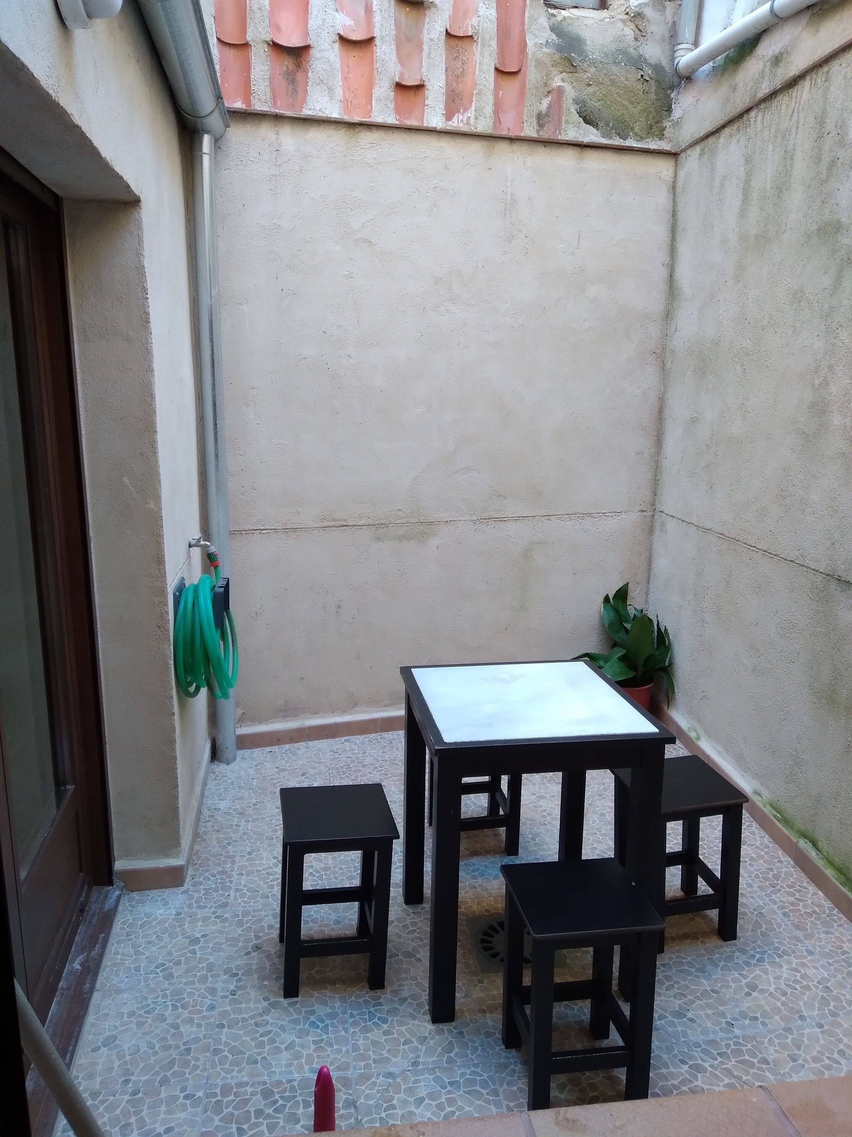 Un pequeño patio exterior con suelo de baldosas, una mesa con cuatro sillas, una planta en maceta y una manguera verde enrollada en la pared.