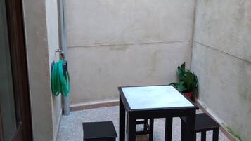 Un pequeño patio exterior con suelo de baldosas, una mesa con cuatro sillas, una planta en maceta y una manguera verde enrollada en la pared.