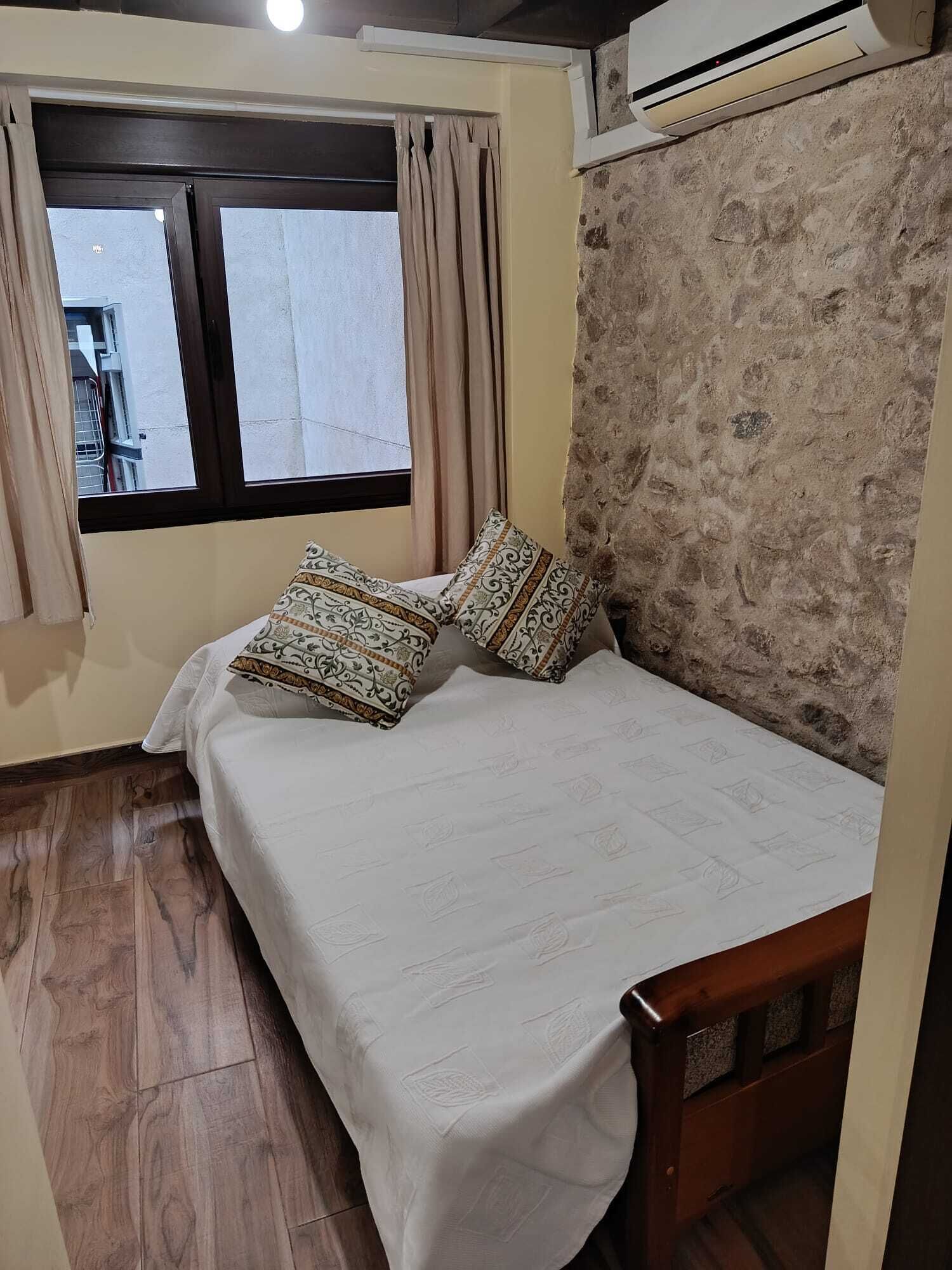A bedroom with a bed, patterned pillows, a window with curtains, a wall with a textured pattern, and an air conditioning unit.
