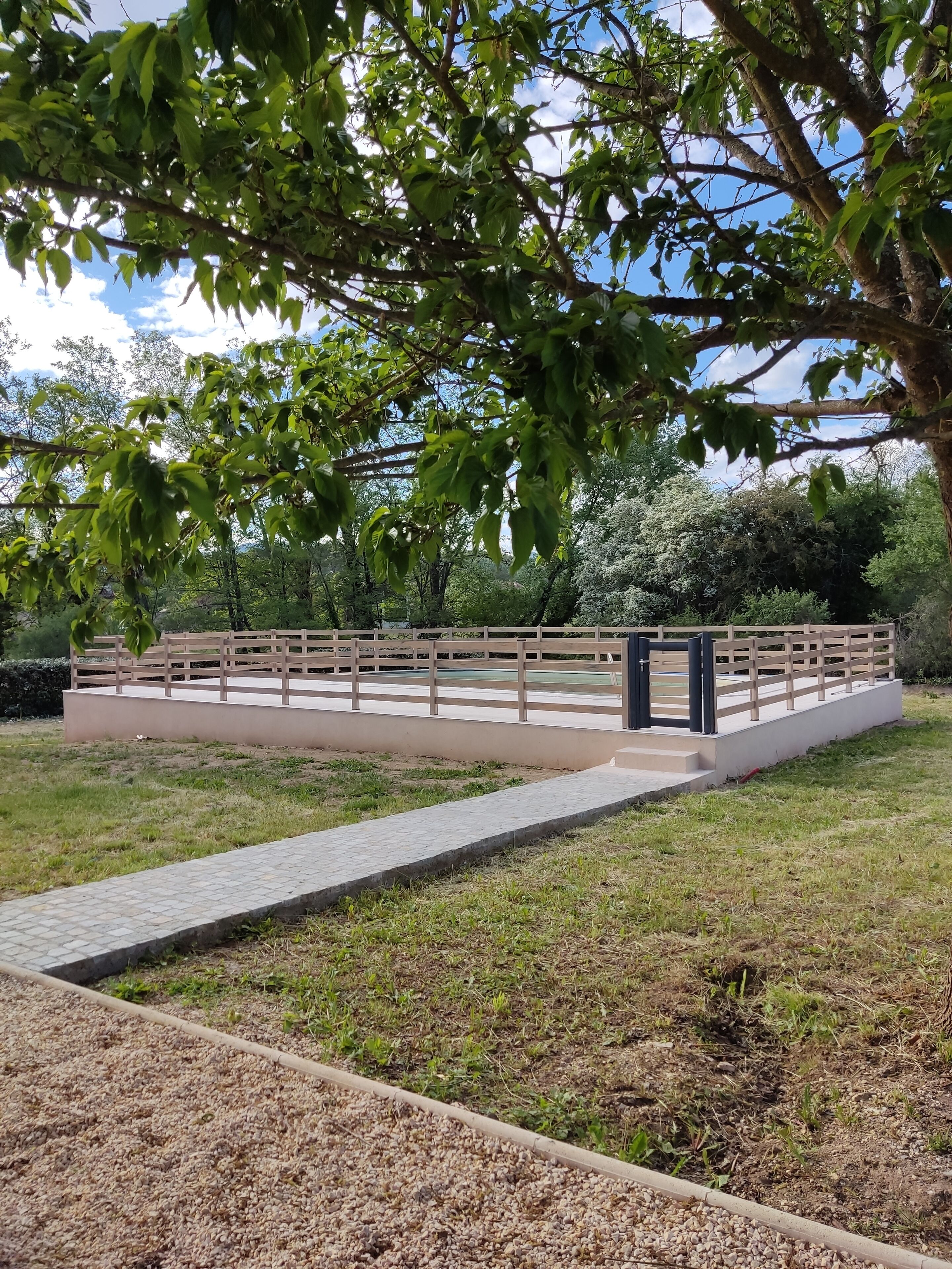 A park with a paved walkway, a wooden fence, and a tree with green leaves.