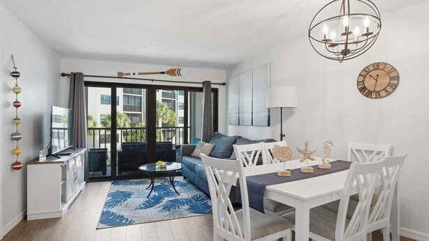 A modern living room with a sofa, dining table, and chairs, a chandelier, and a view through large windows.
