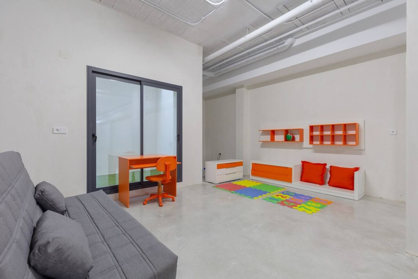 A modern living room with a grey sofa, an orange desk, and a colorful play mat.
