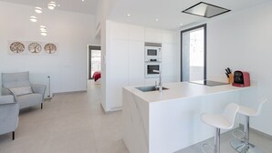 Une cuisine moderne avec des meubles blancs, un îlot central doté d’un évier et d’un four encastré. On y trouve deux tabourets de bar et un appareil électroménager rouge posé sur le plan de travail.