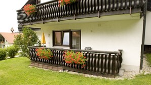A two-story building with a balcony adorned with red flowers, a window with a shutter, and a small garden area.