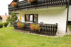 A two-story building with a balcony adorned with red flowers, a window with a shutter, and a small garden area.