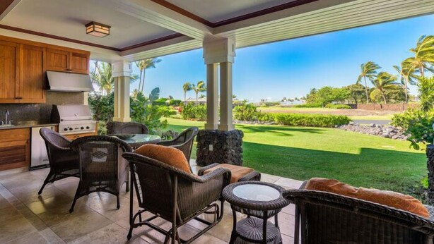 A covered outdoor patio with wicker furniture, a grill, and a view of a well-maintained lawn and palm trees.