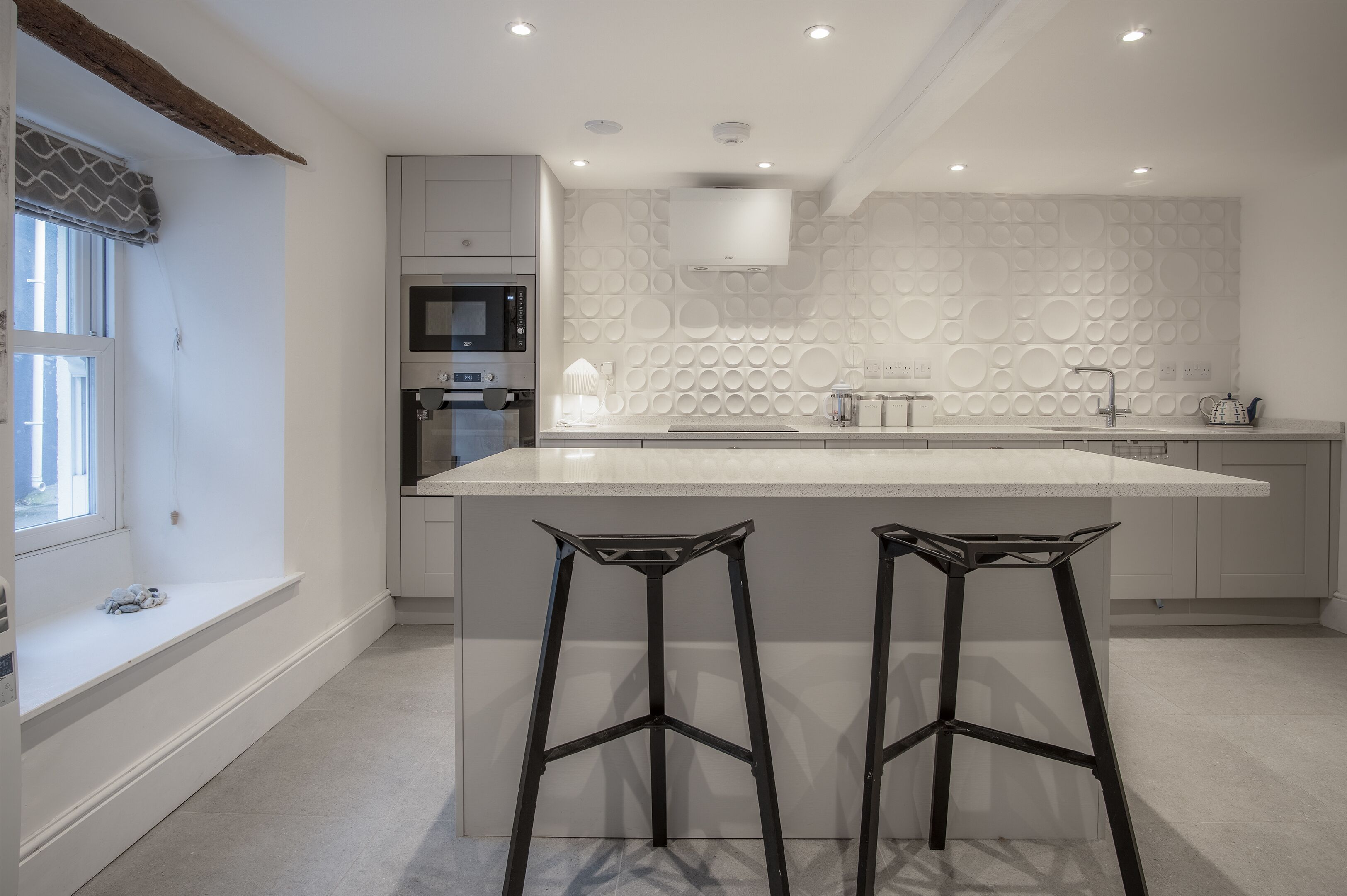 A modern kitchen with a large island, two bar stools, a built-in oven, and a patterned backsplash.