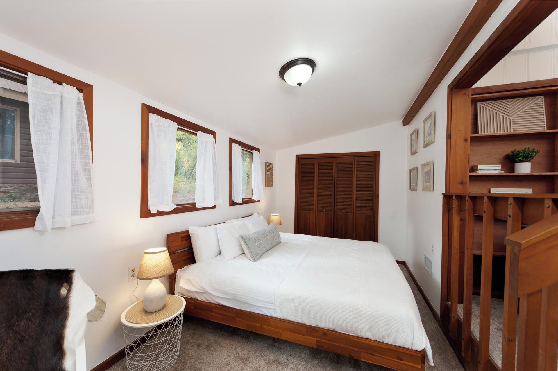 Un dormitorio con cama de madera, ropa de cama blanca, una lámpara de noche y una ventana con cortinas.