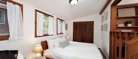 Un dormitorio con cama de madera, ropa de cama blanca, una lámpara de noche y una ventana con cortinas.