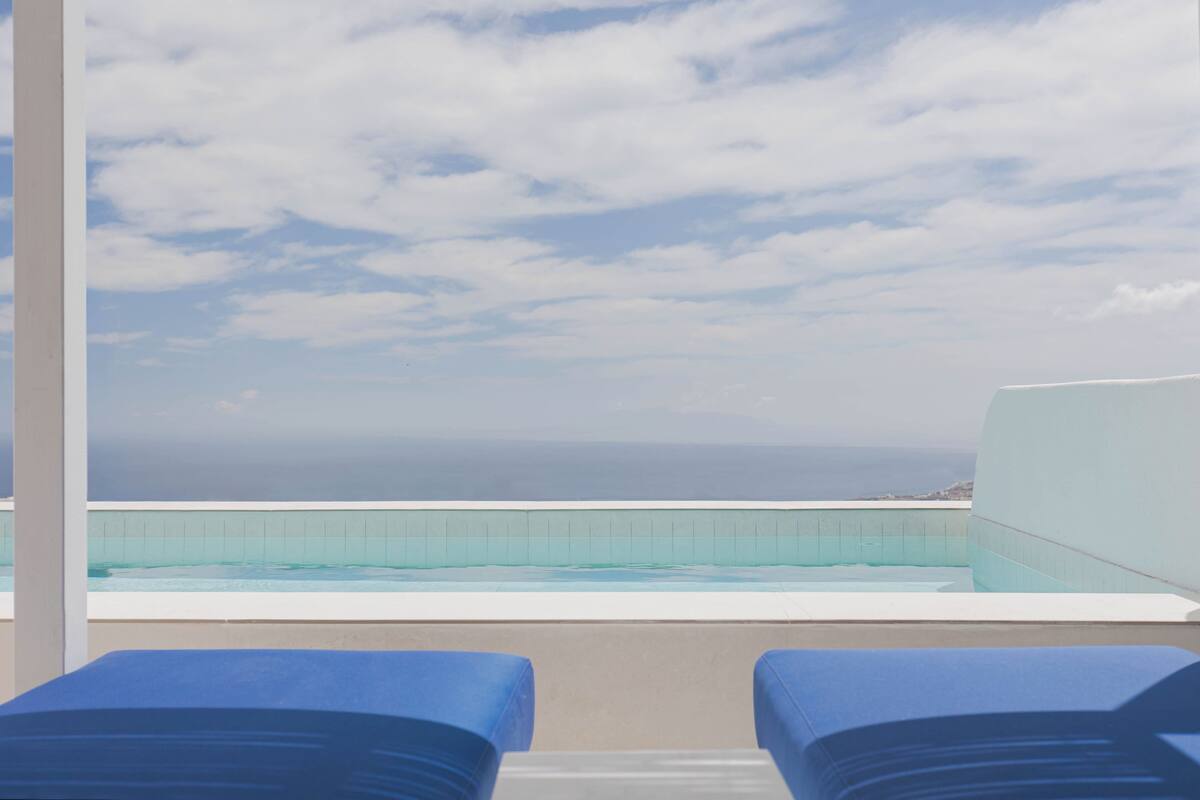 Blick über den Pool mit zwei blauen Liegestühlen, blauem Himmel und weiter Meerblick.