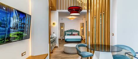 A modern hotel room with a wooden slat wall, a large bed with a blue headboard, a round glass table with blue chairs, and a TV displaying a cityscape.
