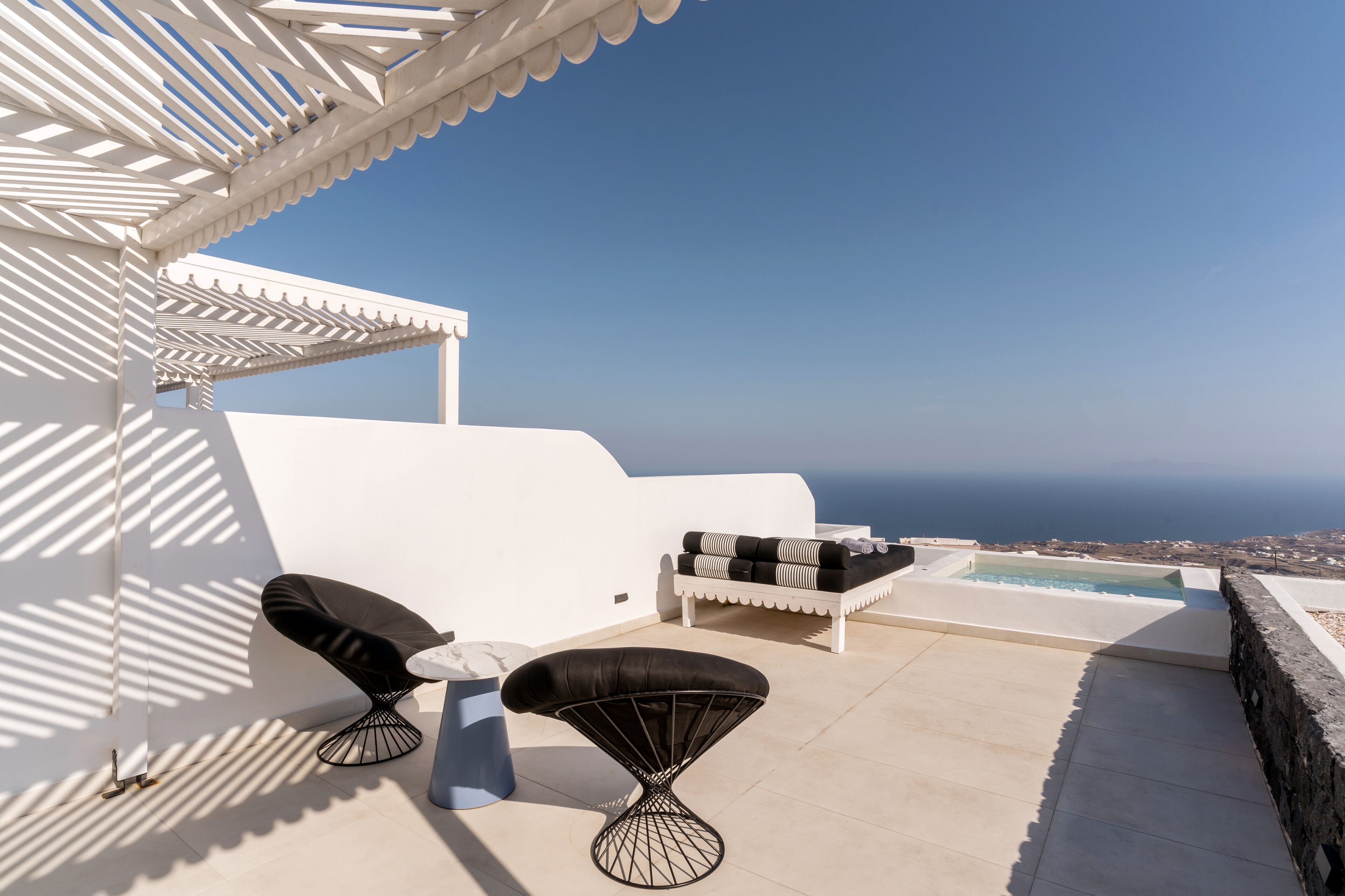 Een terras op het dak met een witte pergola, twee zwarte fauteuils met kussens, een witte bank met zwarte kussens en een zwembad.