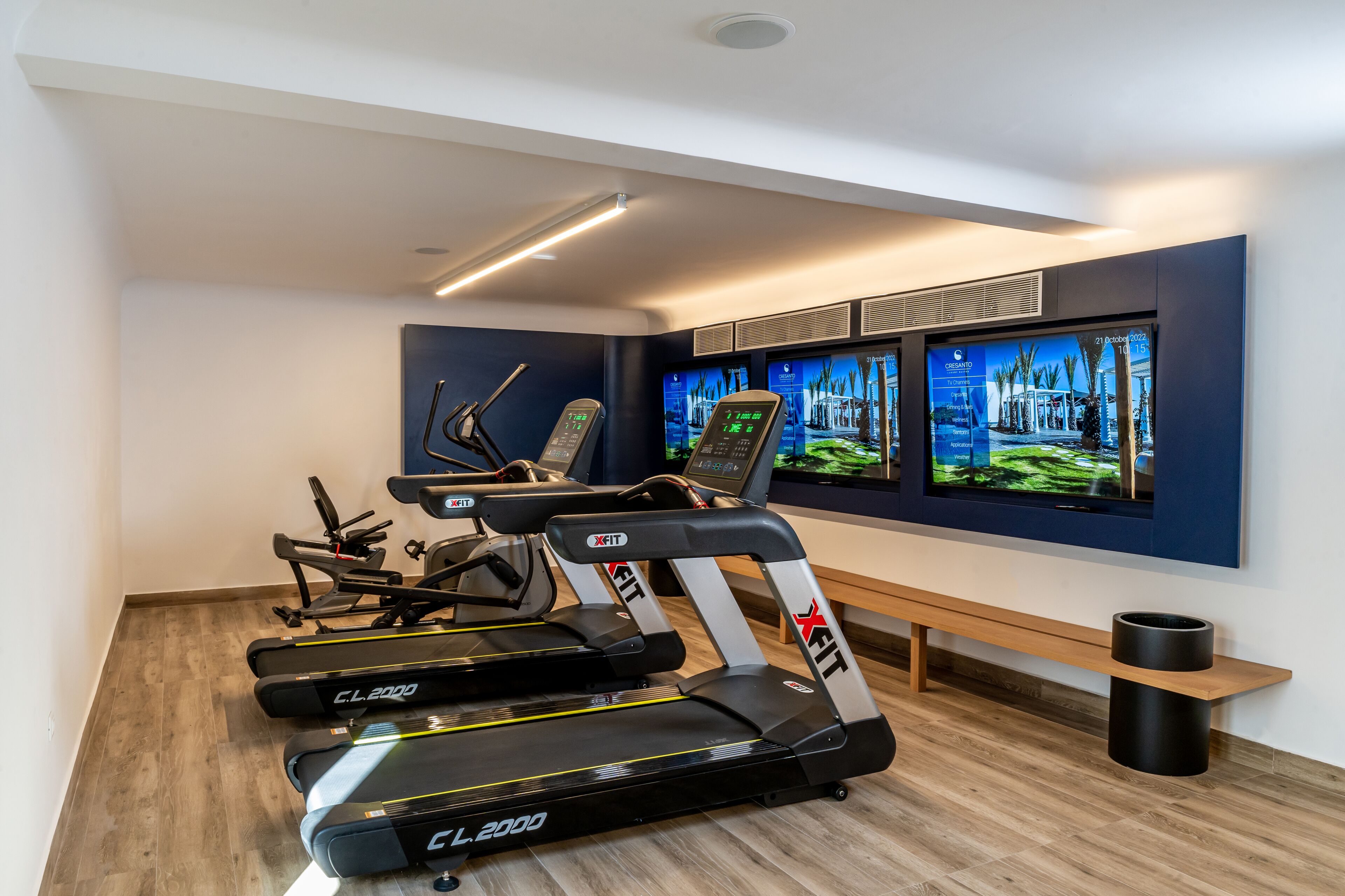 Un gimnàs modern amb cintes de córrer, una banca i un televisor fixat a la paret que mostra contingut sobre fitness.