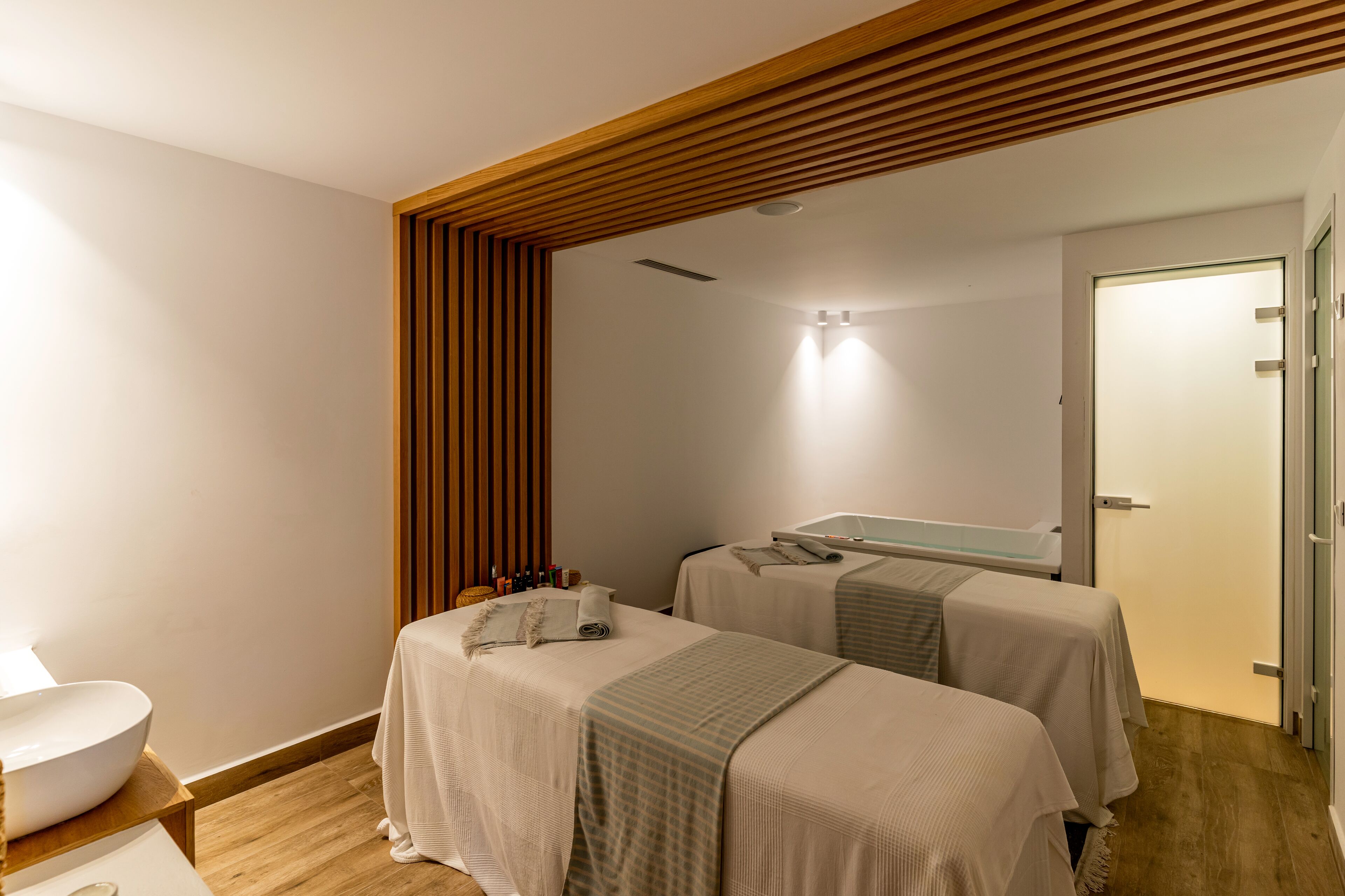 Une chambre d’hôtel moderne avec deux lits, un grand miroir, un plafond en bois et une salle de bain avec un lavabo blanc.